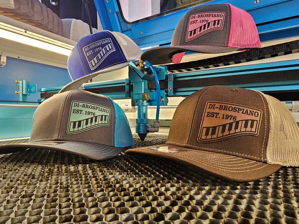 Four custom leatherette patch caps arranged on a work surface near a laser engraving machine, showcasing different colors and designs.