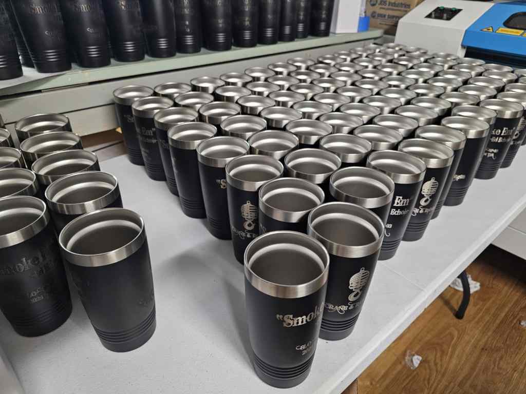 A collection of laser-engraved tumblers lined up on a table, showcasing their black exterior and stainless steel rims.