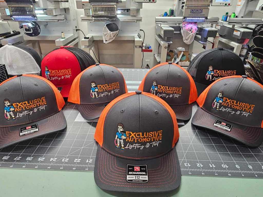 A collection of gray and orange baseball caps featuring an embroidered logo that reads 'EXCLUSIVE AUTOMOTIVE' and an illustration of a person holding tools. The caps are displayed on a work table, with embroidery machines and tools visible in the background.