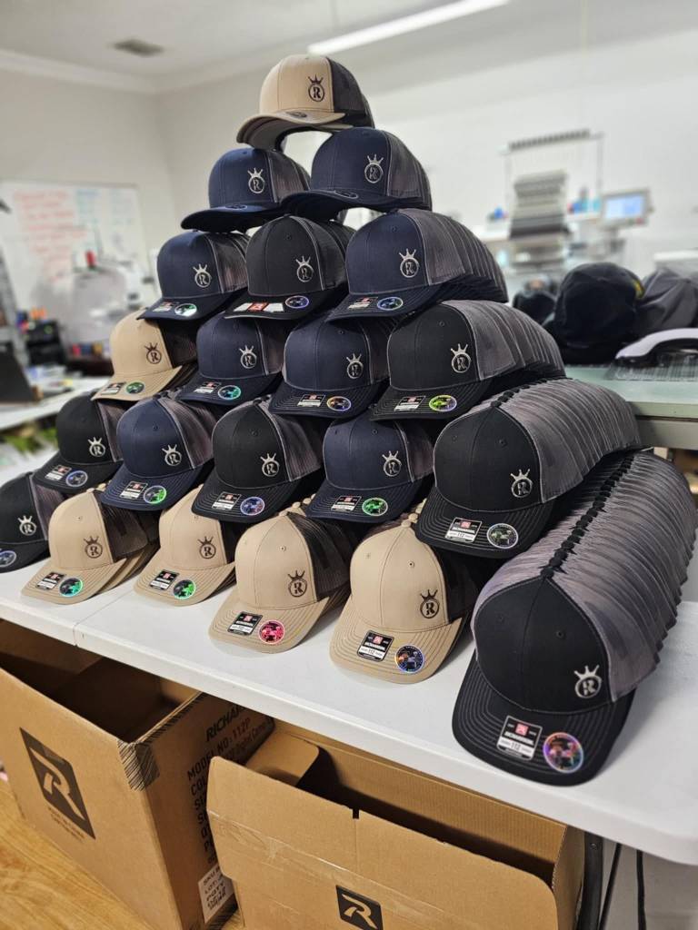 A stack of custom embroidered caps in various colors displayed on a table in a workspace, showcasing their logos.