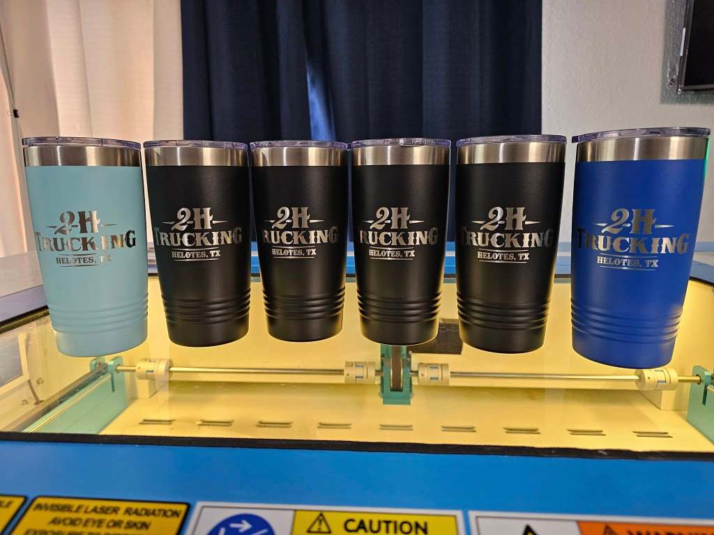 Six laser-engraved tumblers in various colors with '2H TRUCKING HELOTES, TX' text displayed on them, positioned on a laser engraving machine.