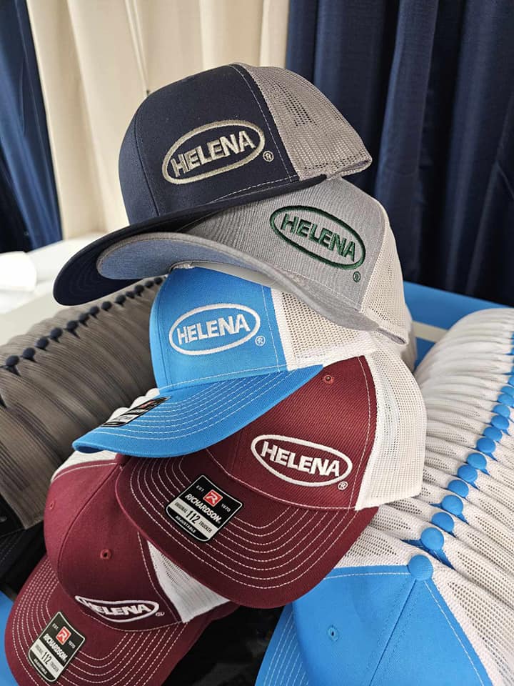 A stack of trucker hats with the brand name 'HELENA' prominently featured on the front, showcasing various colors including navy, green, blue, and maroon.
