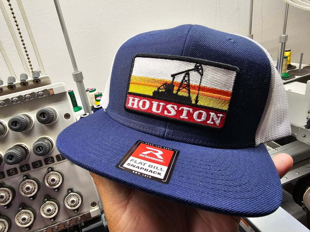 A close-up of a navy blue cap with a flat bill, featuring an embroidered patch that reads 'HOUSTON' with an oil rig graphic against a sunset background. The cap is being held by a hand in front of a machine.