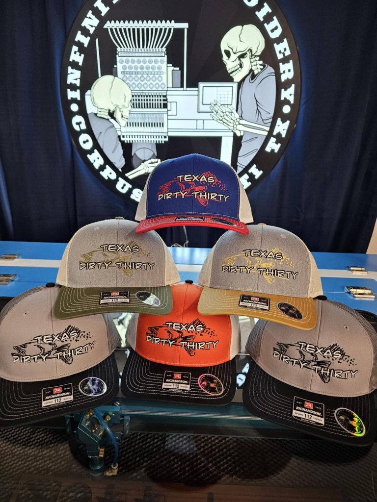 A display of various caps with 'Texas Dirty Thirty' embroidered on the front, arranged in front of an embroidery machine backdrop.