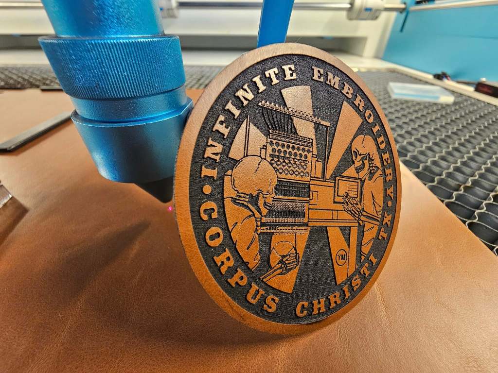 A close-up of a laser engraving machine working on a circular leather patch featuring the logo and design of Infinite Embroidery, showcasing a creative illustration of two figures and text.